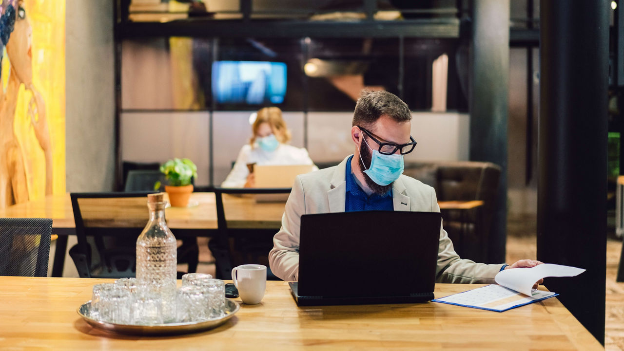 A man wearing a face mask sits at a table with a laptop.