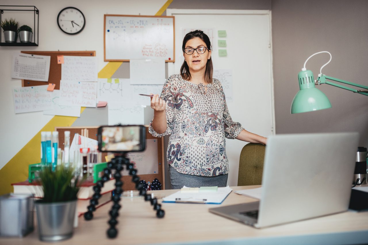 Woman holding a pen and standing behind a desk records herself with a phone on a tripod.