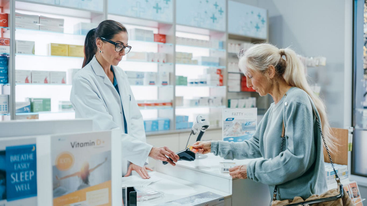 Pharmacist assists an older woman paying at a pharmacy counter with a card reader, with shelves of medicines and health products in the background.
