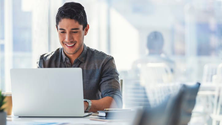 A man is smiling while working on a laptop in an office.