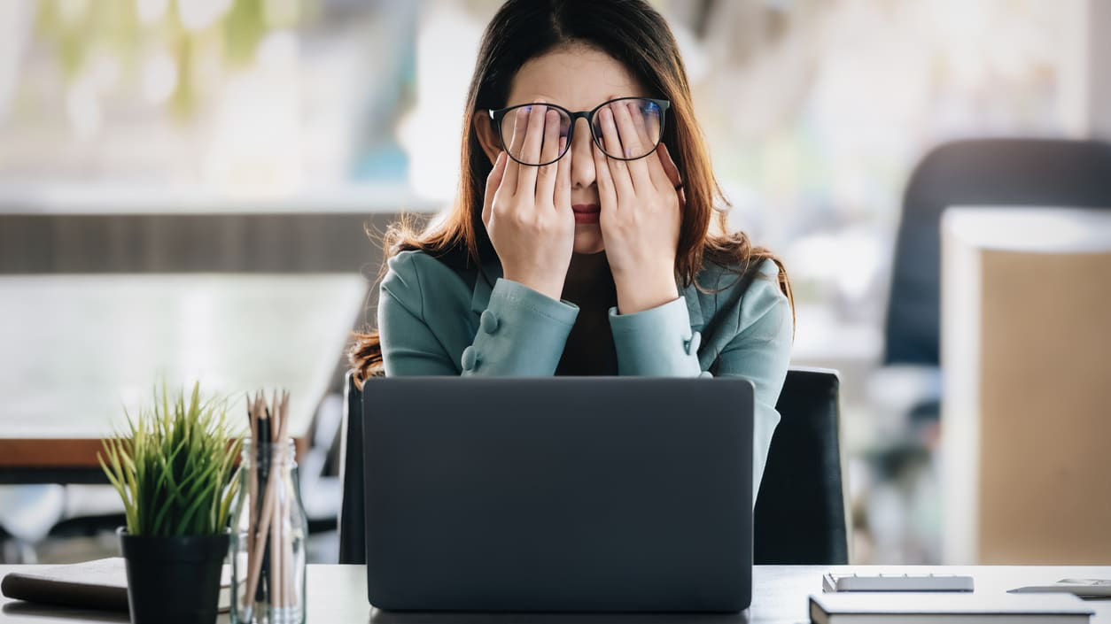 A woman covering her eyes while working on a laptop.