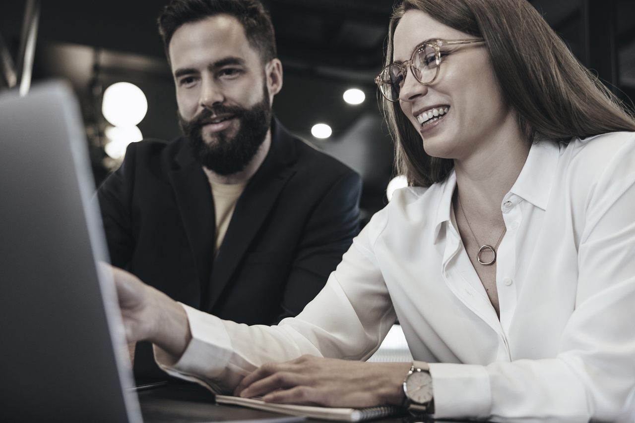 Young businesspeople smiling while using a laptop in an office. Happy young entrepreneurs collaborating in a modern workspace. Two successful businesspeople sitting together at a table.