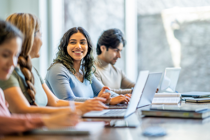 A small group of four mature adult students sit together at a long desk as they study with one another. They have text books and laptops out in front of them as they take notes and talk amongst themselves.