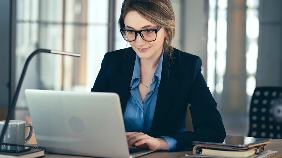 A woman in glasses is working on a laptop in an office.