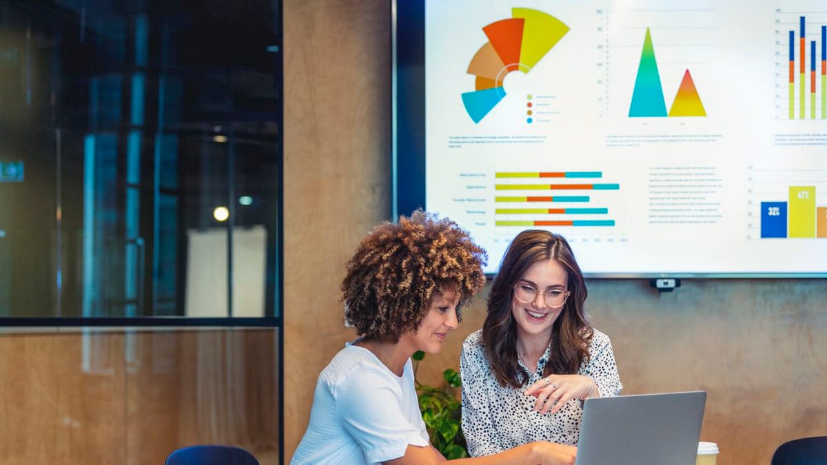 Two women working on a laptop in a conference room.