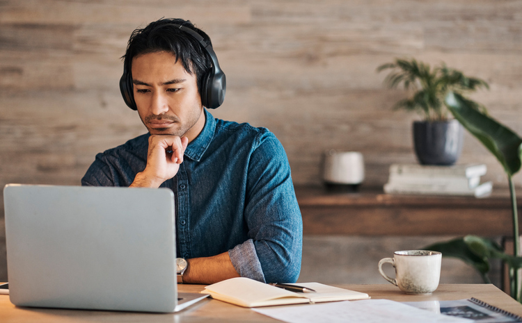 Businessman, headphones and laptop webinar in office with coffee on table, video call or watching video. Zoom call, video conference and male from Canada in online meeting with book and pen on desk.