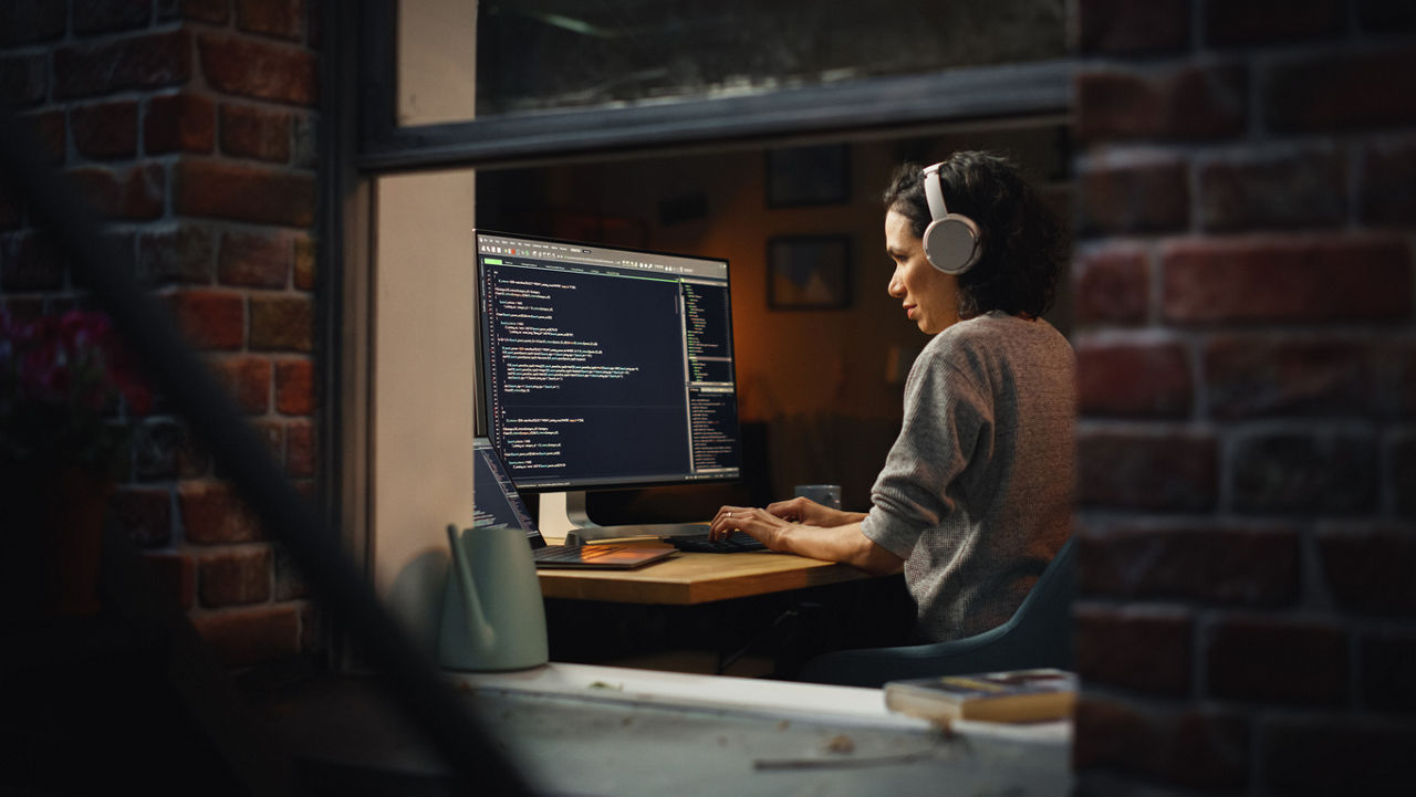 Hispanic Female Programmer in Headphones Listening Podcast and Working on Desktop Computer, Screen Shows Coding Language User Interface. Digital Entrepreneur Creating Modern Software.