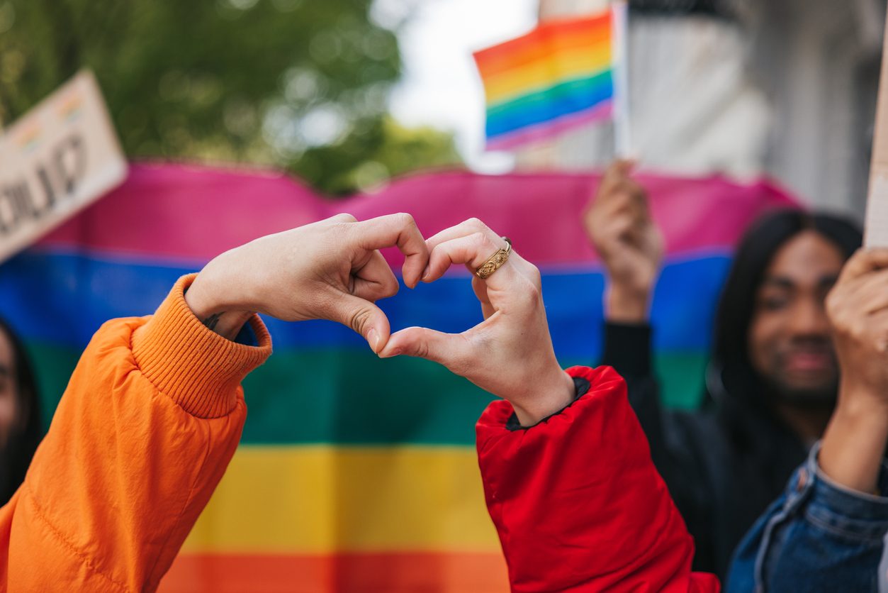 hand makes heart with Pride flag in background