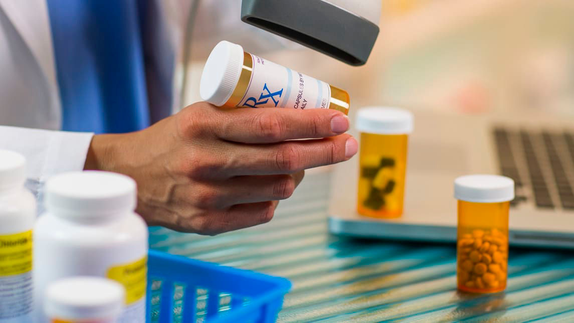 A pharmacist is holding a bottle of medicine in front of a laptop.