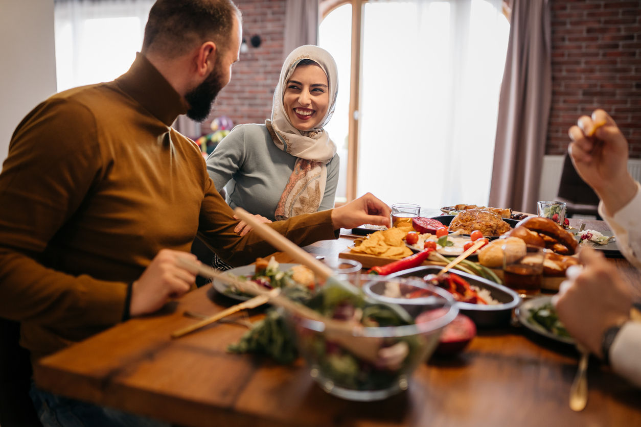 Group of adults seated around a dining table sharing a meal, smiling and talking, with a woman wearing a headscarf at the center of the gathering.