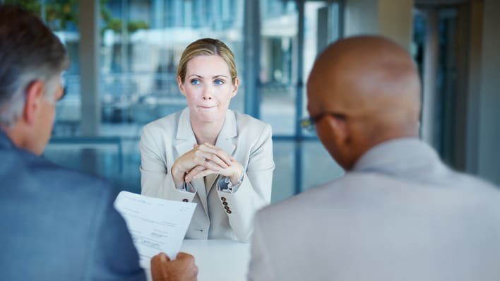 Two interviewers sit across from a female candidate, who appears slightly nervous or uncertain. The candidate is seated with her hands clasped in front of her, while one interviewer holds a document, possibly her resume. The setting is a professional, modern office space with large windows in the background.