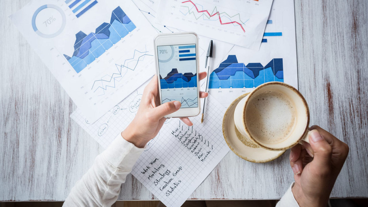 A woman is holding a smartphone while looking at graphs on a table.