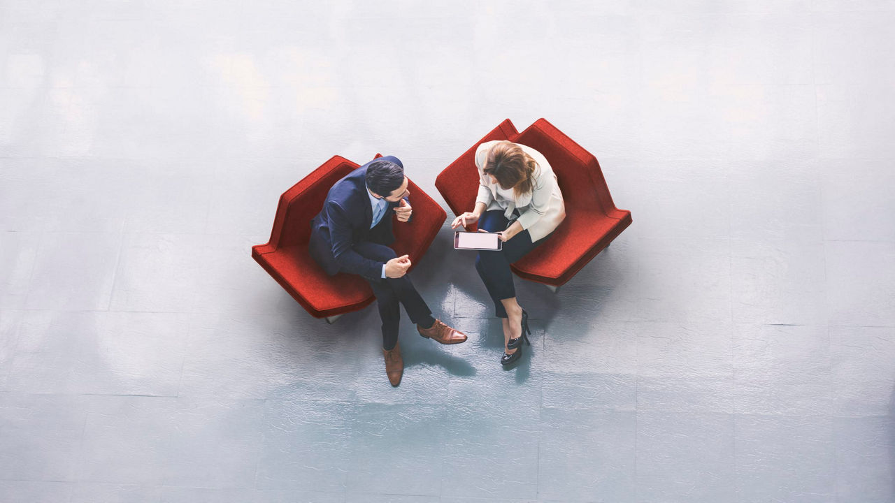 Two business people sitting on red chairs in an office.