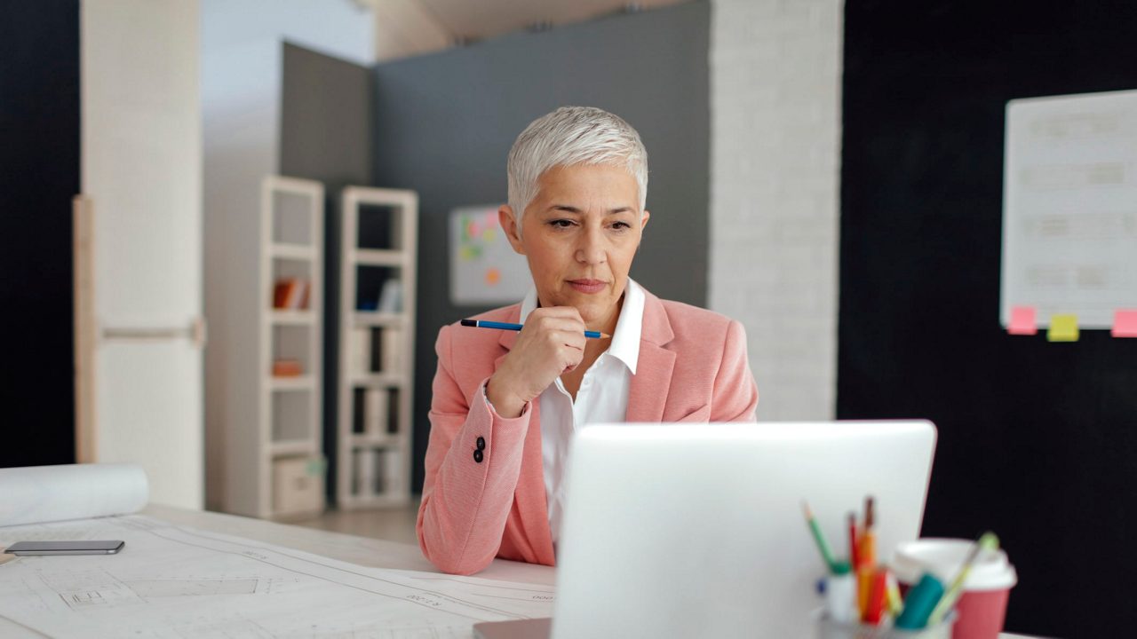 A woman is sitting at a desk looking at a laptop.