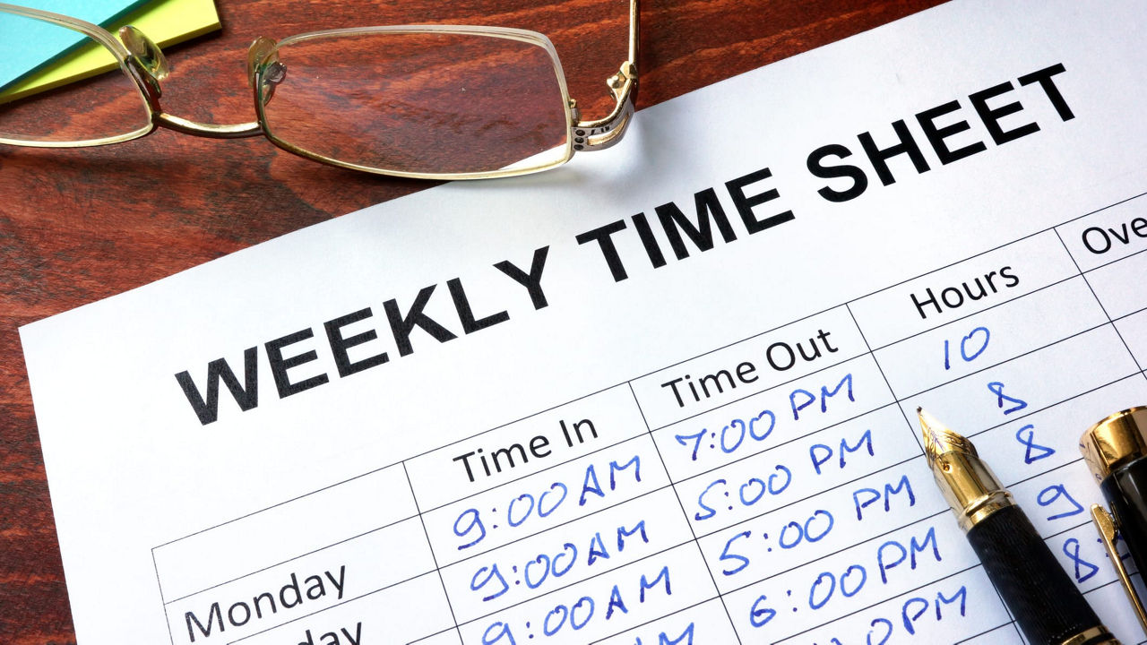 A weekly time sheet with glasses and pens on a wooden table.