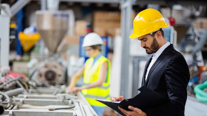 A man in a hard hat is holding a clipboard in a factory.
