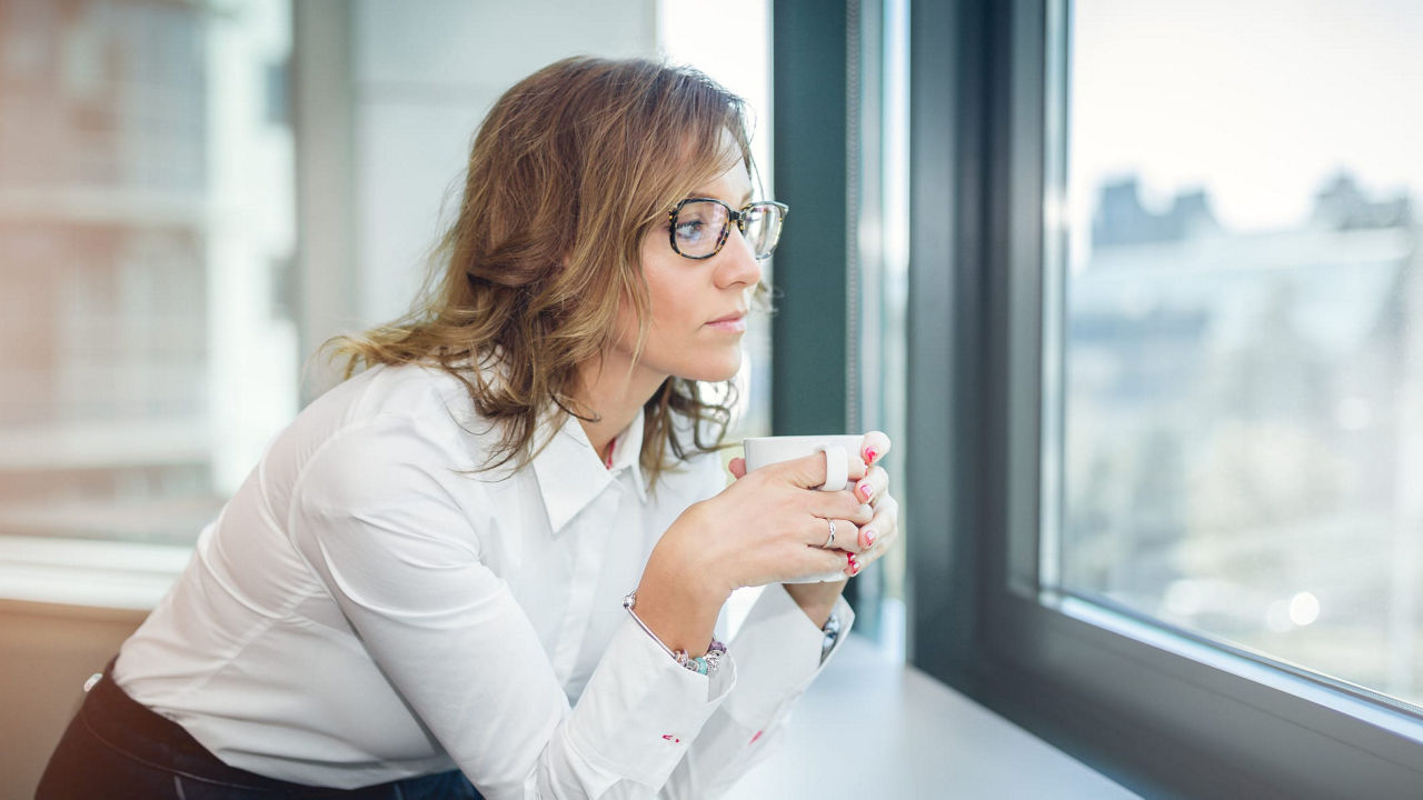 A woman is leaning against a window sill and drinking a cup of coffee.