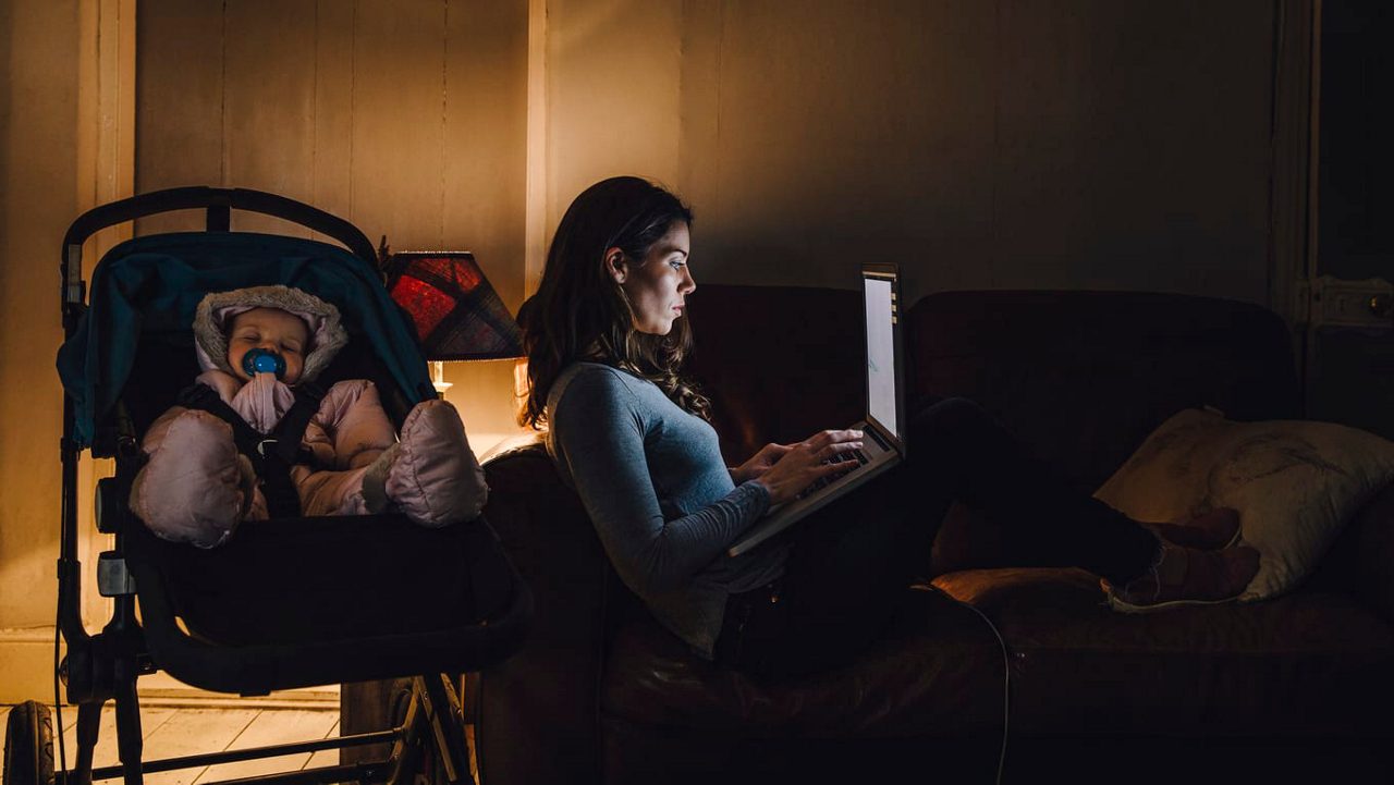 A woman sitting on a couch with a baby in a stroller and a laptop.