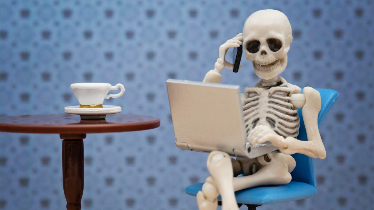 A skeleton sitting in a chair with a laptop and a cup of coffee.