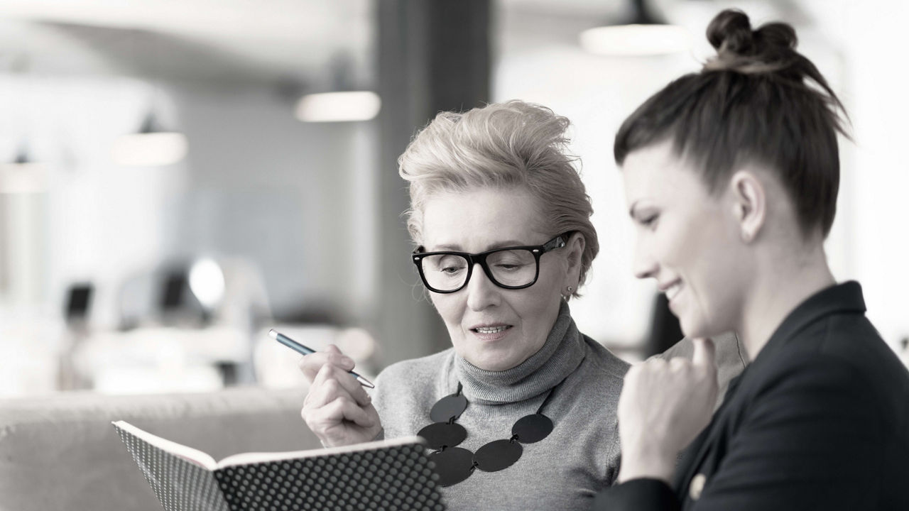 Two women in glasses are looking at a notebook.