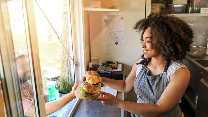 A woman in an apron is holding a sandwich out of a window.
