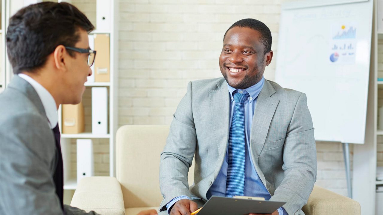 Two businessmen sitting in an office talking to each other.