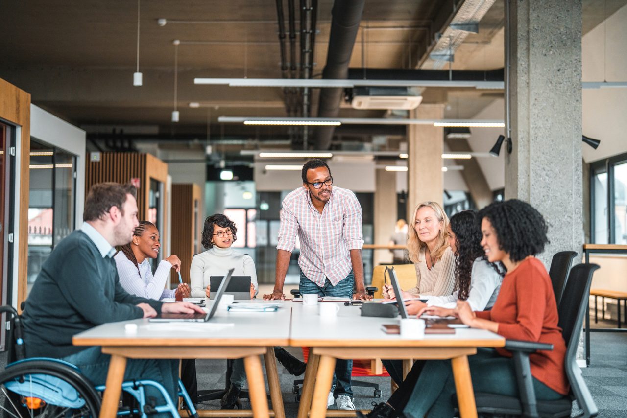 Bright open concept office. There is a work meeting being held where a group of diverse people, including a man on a wheelchair are working together.