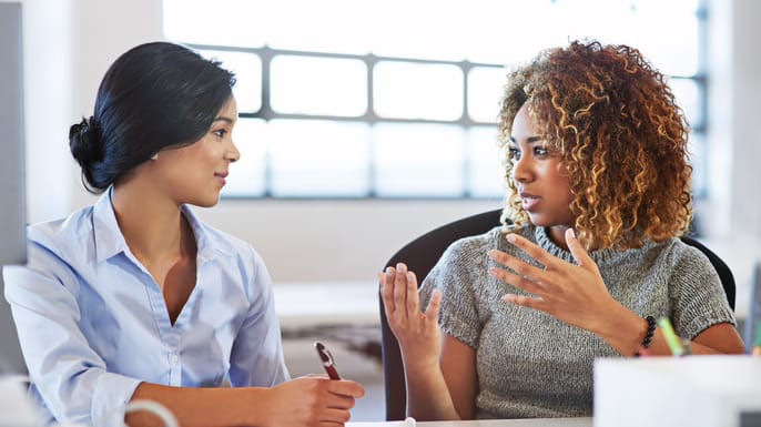 Two women talking at a desk in an office.