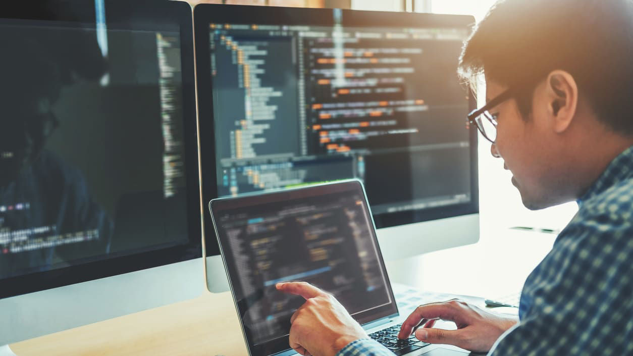 A man is working on a laptop in front of two monitors.