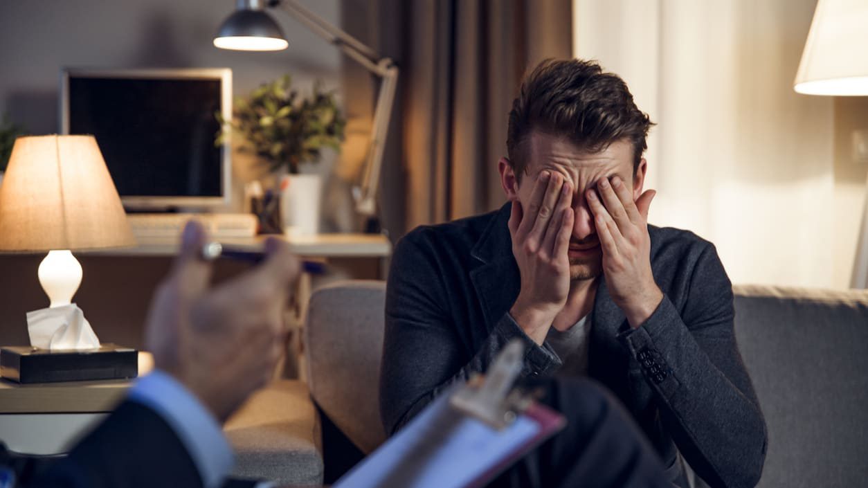 A man is sitting on a couch with his hands covering his eyes.