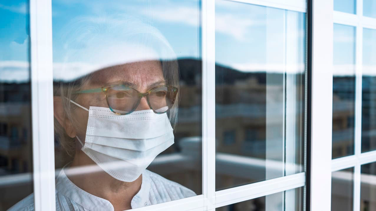 A woman wearing a surgical mask looking out of a window.
