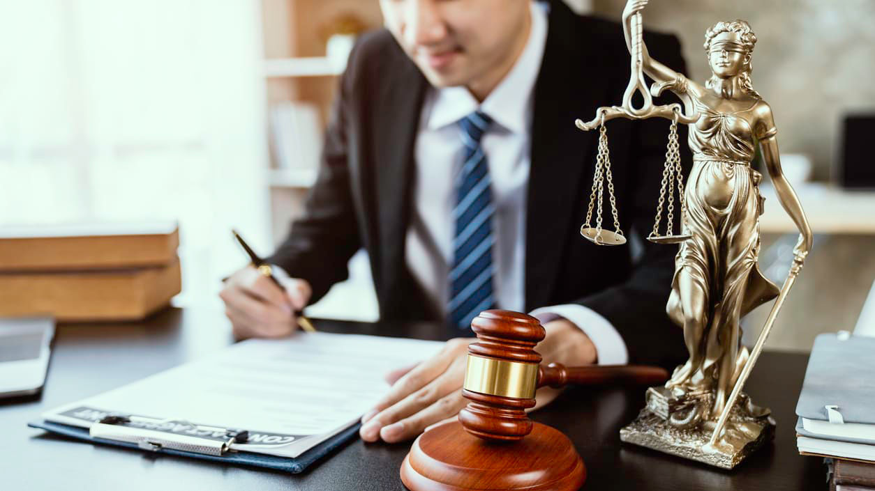 A man in a suit sitting at a desk with a statue of justice.