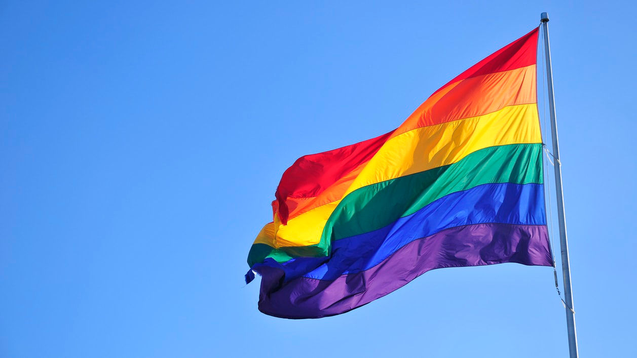 A rainbow flag flying in the wind against a blue sky.