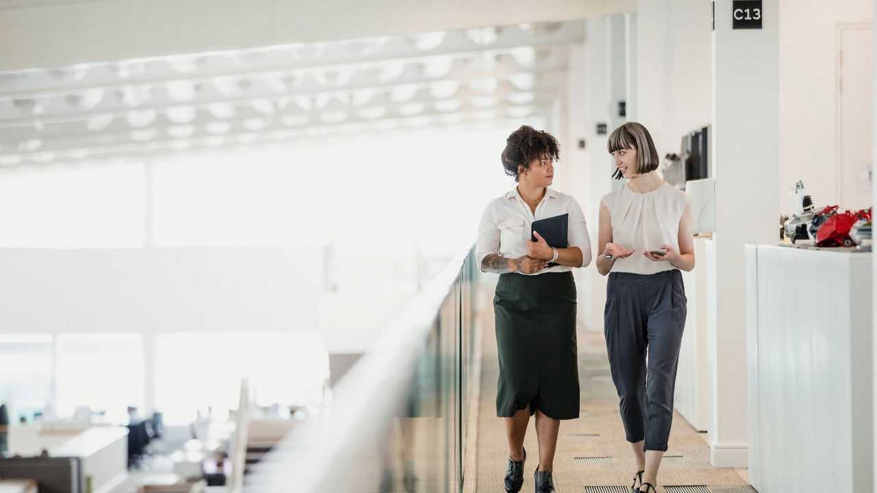 Two business women walking down a hallway in an office.