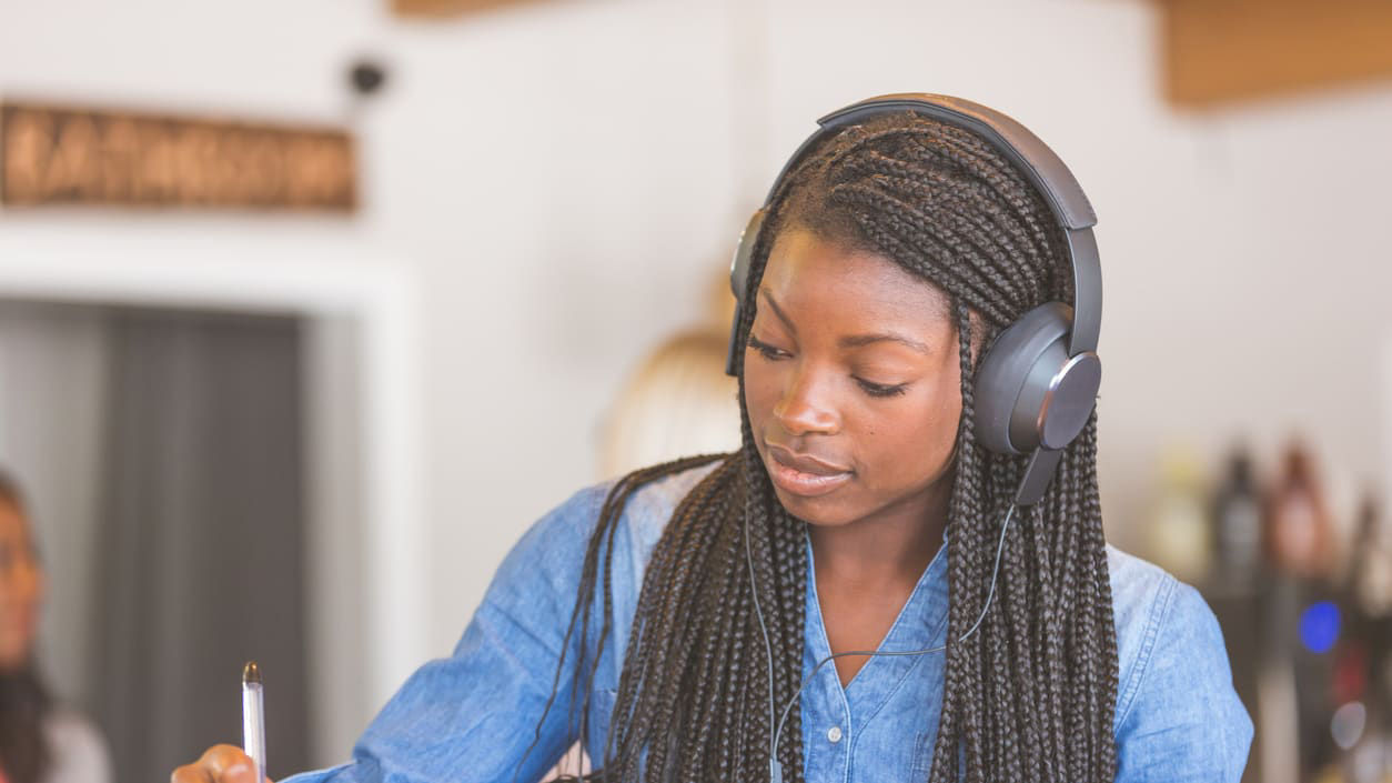 A woman wearing headphones is writing in a notebook at a coffee shop.