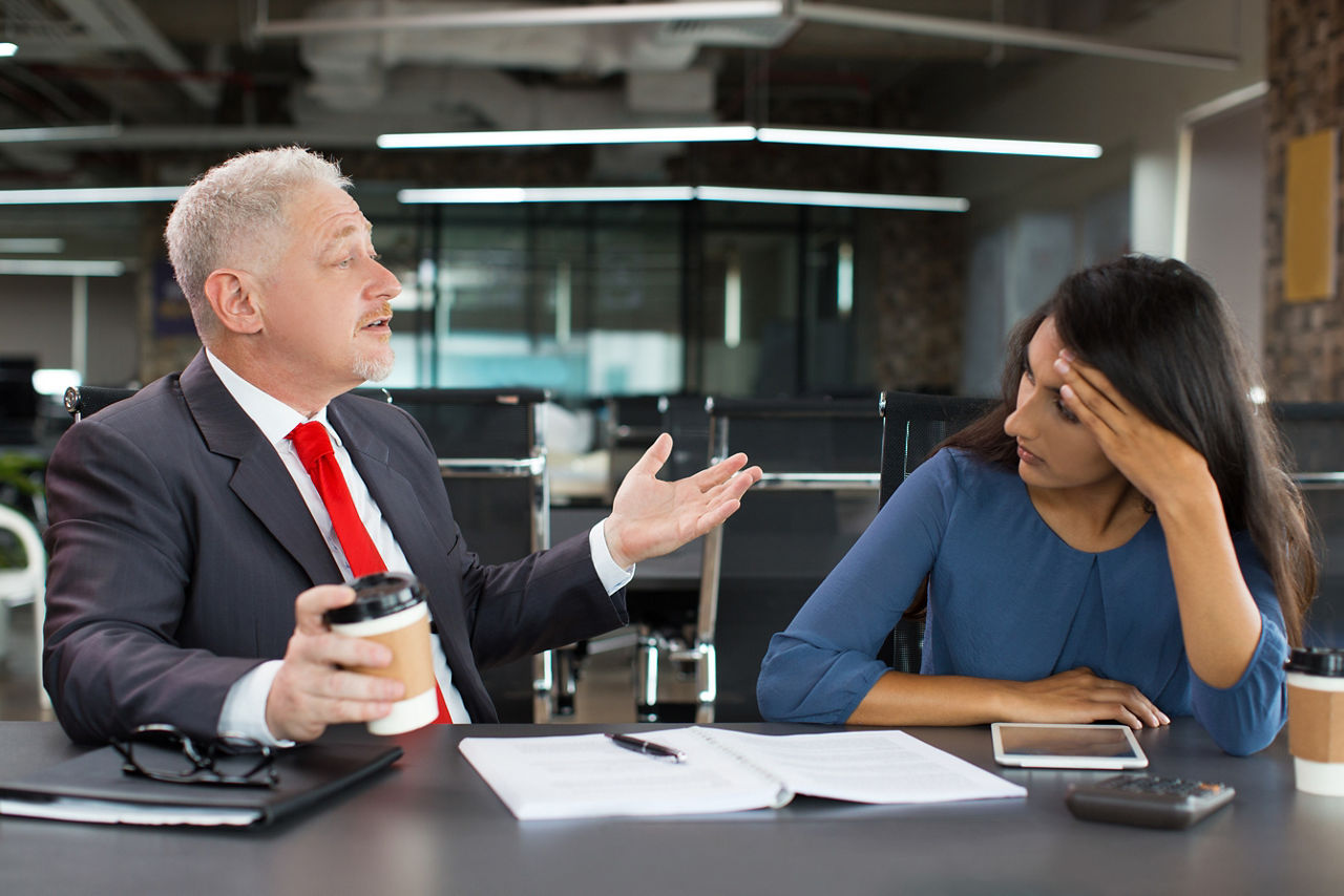 Portrait of tired or bored young Indian businesswoman sitting at table and listening to her senior Caucasian colleague or mentor in factory office. Business relationship and internship concept
