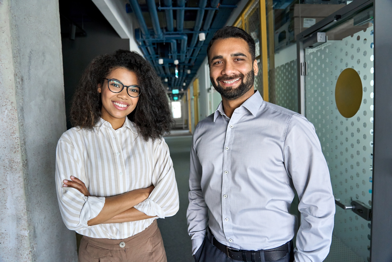 Two happy diverse professional executive business team people African American woman and Indian man looking at camera standing in office lobby hall. Multicultural company managers team portrait.