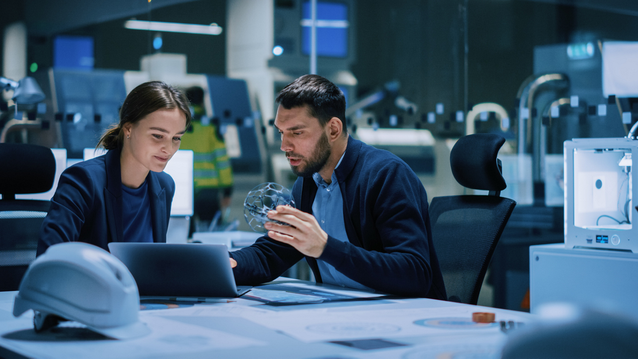 Industry 4.0 Modern Factory Meeting Room: Chief Engineer Holds Mechanism, Shows it to Female Designer, They Talk, Use Laptop. Scientists in Lab Build Electronic Machinery for With Futuristic Design