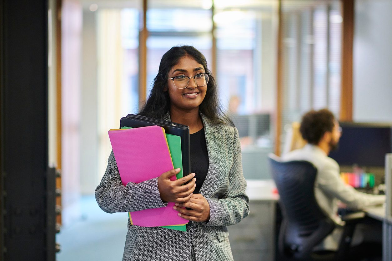 Young woman wearing glasses and a blazer smiles and holds multicolored folders in an office setting