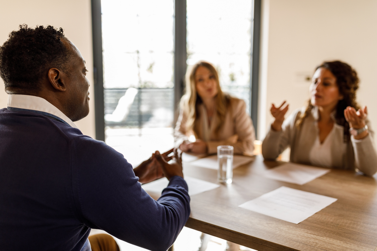 Over the shoulder view of confident man sitting at table, his resume in front of him, talking about his skills and education during a job interview meeting with employers sitting opposite him.