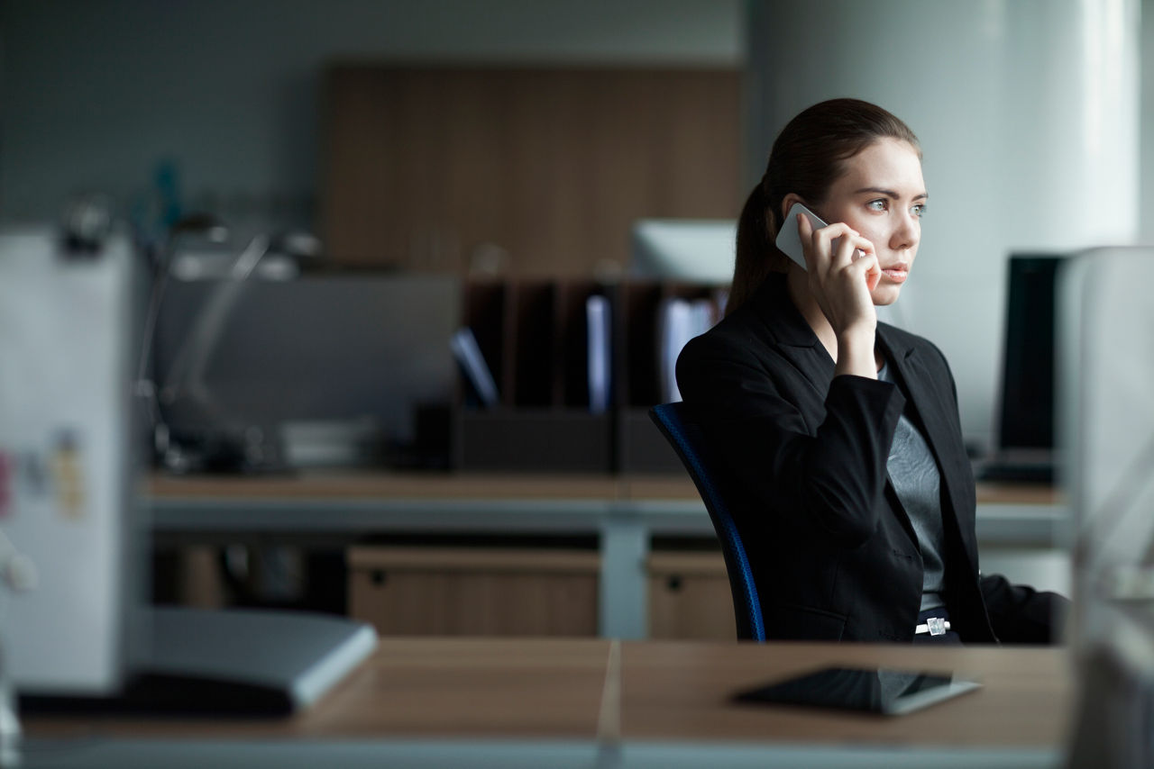 Professional woman in a business suit sitting at a desk in a dimly lit office, attentively listening while speaking on the phone, conveying a serious and focused expression.