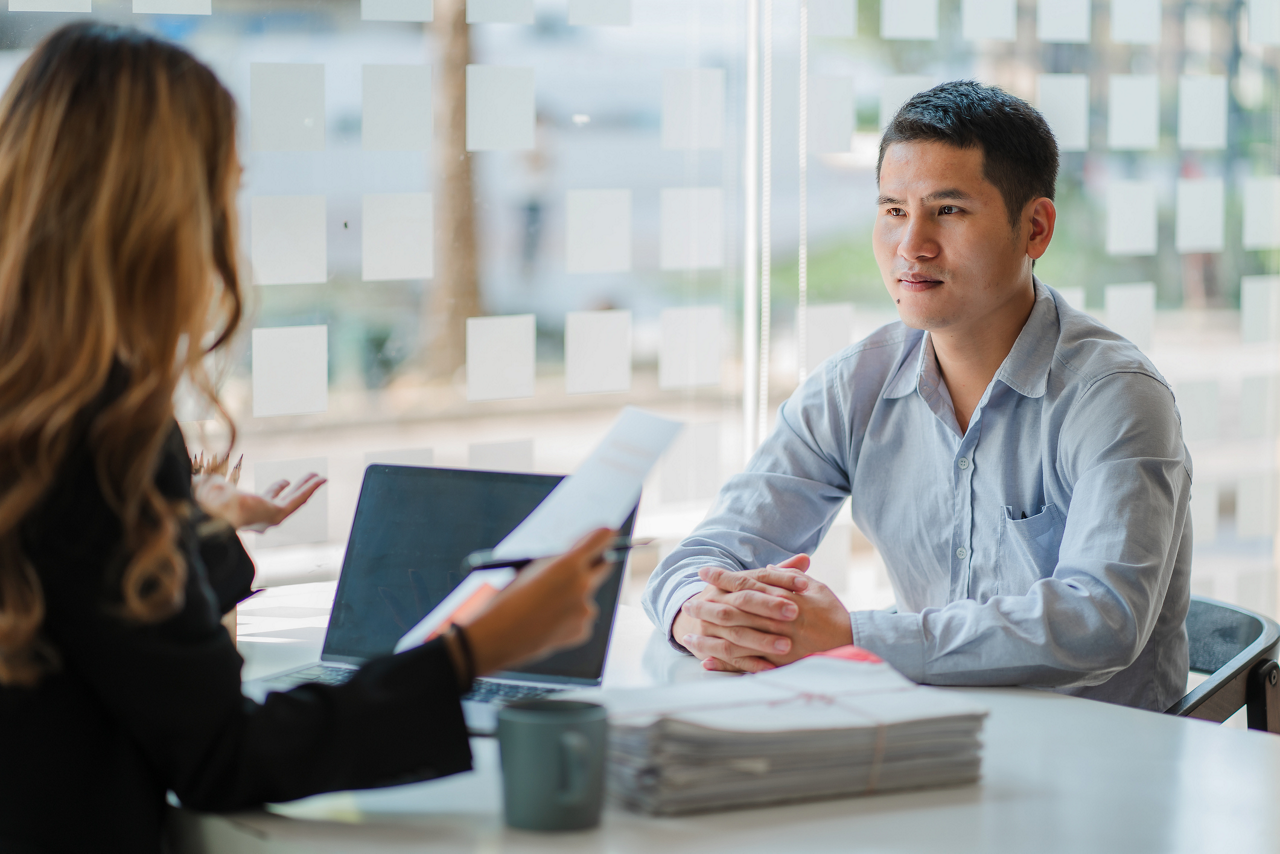Job candidate sits across desk from woman interviewing him, her back is turned to camera