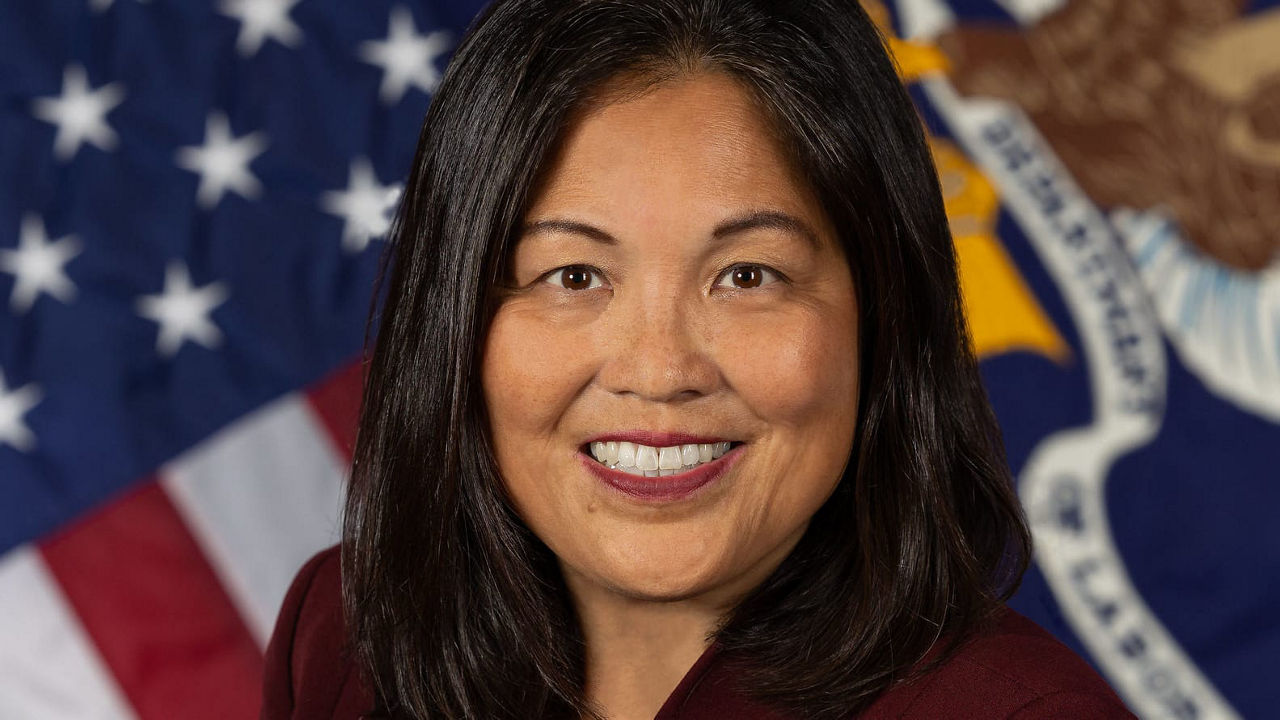 A woman in a maroon jacket standing in front of an american flag.