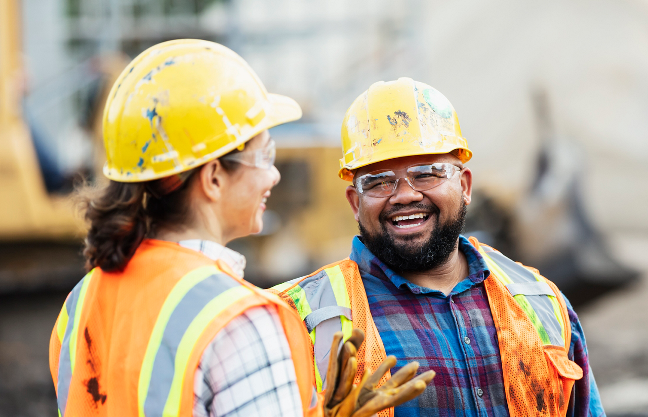A group of three multi-ethnic workers at a construction site wearing hard hats, safety glasses and reflective clothing, smiling and conversing. The main focus is on the mixed race African-American and Pacific Islander man in the middle. The other two construction workers, including the woman, are Hispanic.