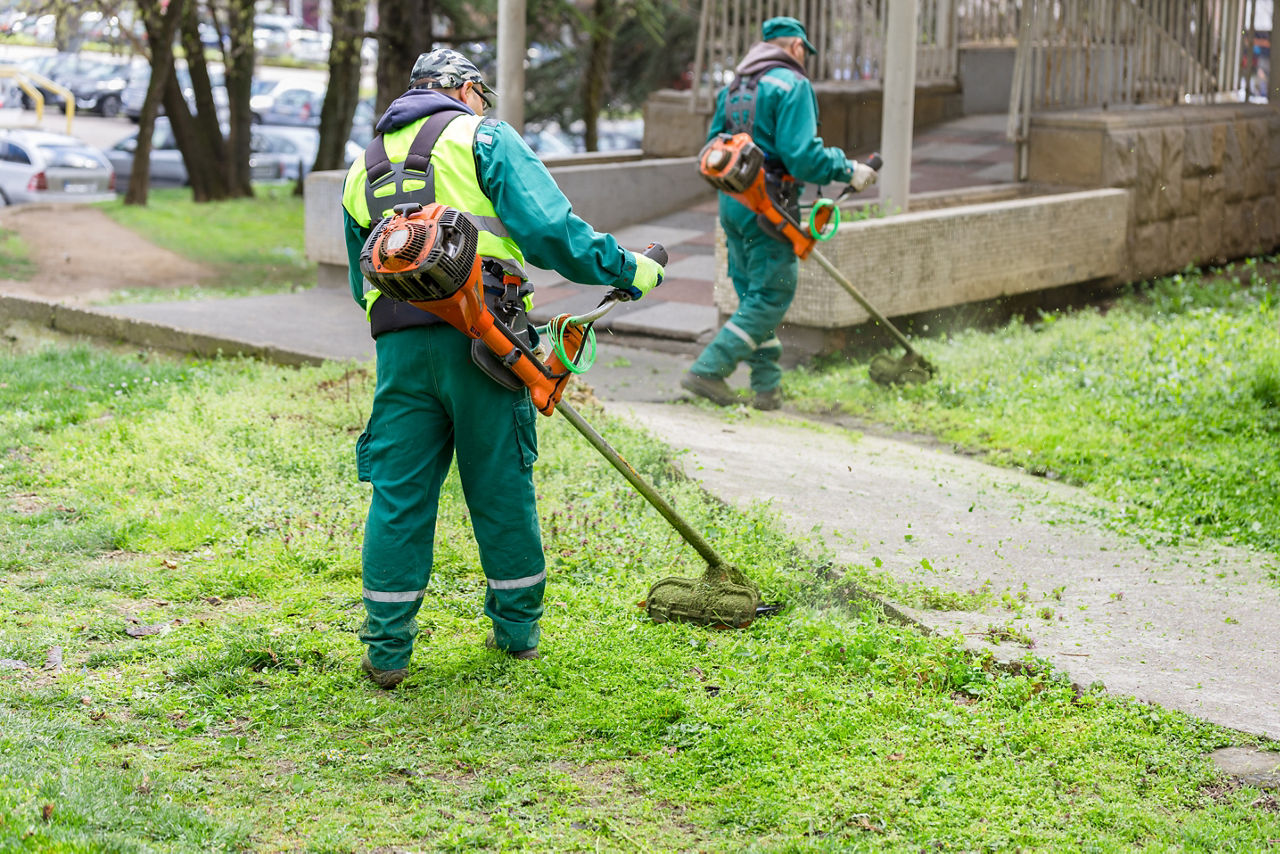 Two workers in green uniforms with safety vests mowing the grass in a city environment, maintaining the urban green spaces for aesthetic looks and safety