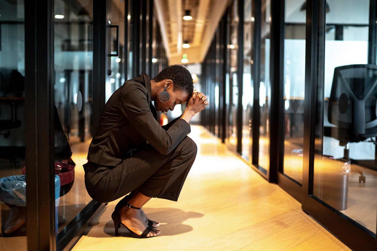 Worried young businesswoman crouching down with head in hands in an office building corridor