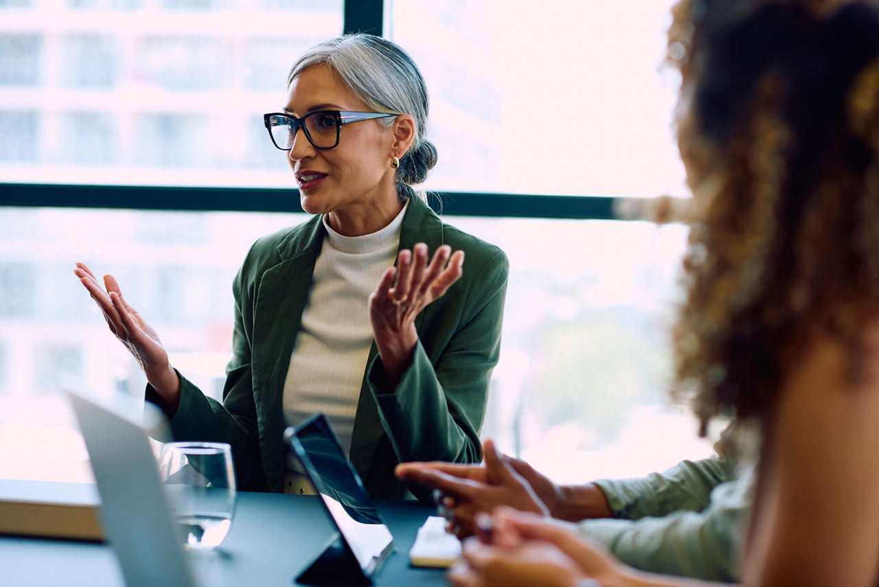 Mature professional woman gesturing while leading a discussion during a business meeting beside digital devices.