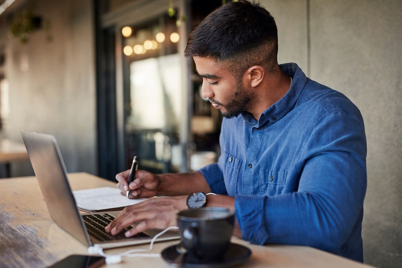 A businessman writes with his right hand as he works on his company's learning and development in a coffee shop or restaurant. He has his left hand on his laptop.