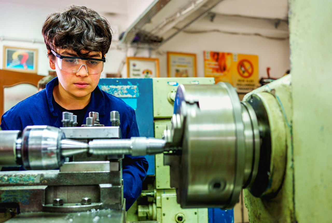 A young male trainee in safety glasses operates a Lathe machine in a technical training workshop. Concept of vocational education, industrial skills development, and hands-on learning in mechanical engineering.