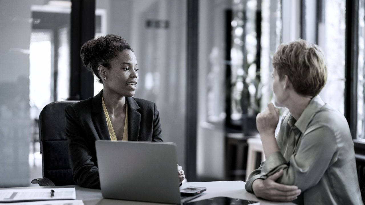 Manager on left speaking to employee on right, both sitting at table in office